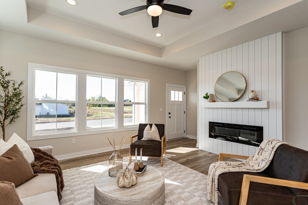 A modern living room with large windows, neutral decor, a round mirror above a fireplace, and three chairs arranged around a round coffee table.