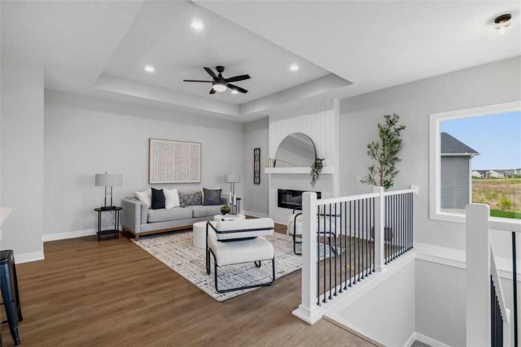 Spacious living room with tray ceiling, black ceiling fan, shiplap fireplace, and open staircase railing in Waukee Iowa new construction
