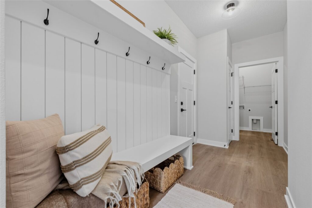 Mudroom with white shiplap wall, black coat hooks, built-in bench, and view to laundry room in Waukee Iowa new construction home