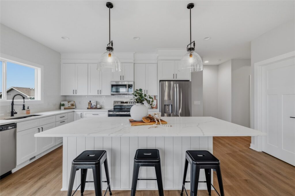 White shaker kitchen with large quartz island, black metal bar stools, glass pendant lights, and stainless appliances in Waukee Iowa new home