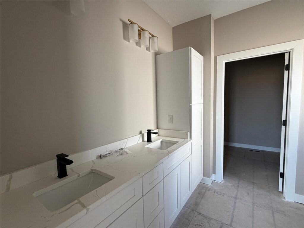 Modern bathroom with double white marble sinks, black faucets, tall storage cabinet, and beige tiled floor leading to a dark doorway.