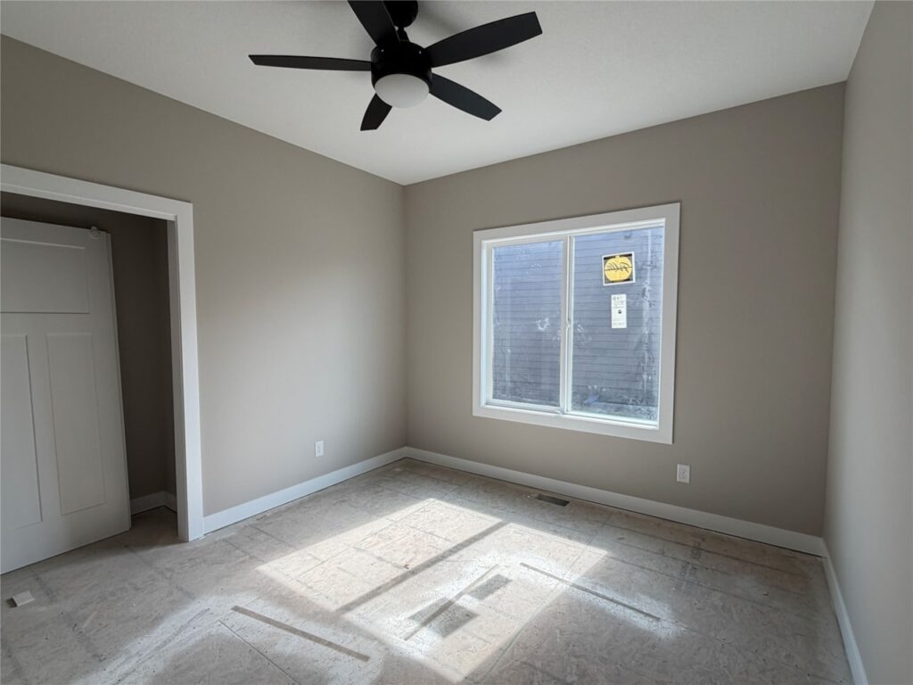 Empty room with beige walls, a ceiling fan, a window casting light on an unfinished floor and an open closet door.