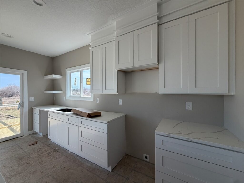 A modern kitchen with white shaker cabinets, light gray walls, and white countertops. It features a black sink under a double-hung window, two floating corner shelves, and a glass-paneled door leading outside. Upper cabinets line the right wall above base cabinets with drawers. The ceiling has recessed lights, electrical outlets are visible, and the floor is unfinished with few appliances, indicating new construction.