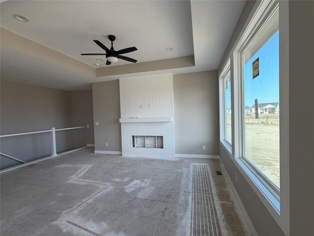Empty modern living room under construction with a white fireplace, large windows, a ceiling fan, and a bare concrete floor.