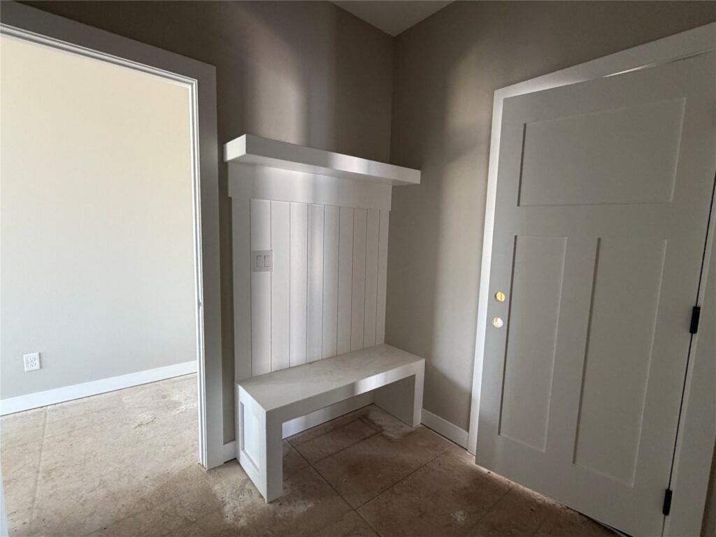 Entryway with a white built-in bench and shelf, beige walls, and a closed gray door, with an open doorway leading to an adjacent room.